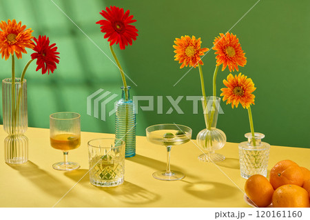 A modern Tet concept in the still life photo template, front view of water glasses placed next to flower vases arranged with gerbera, and tangerines dish on a yellow table, in the green background. A modern Tet concept in the still life photo template, front view of water glasses placed next to flower vases arranged with gerbera, and tangerines dish on a yellow table, in the green background. 120161160