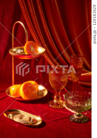 A close-up New Years Eve party idea image has a variety of glasses next to a brass tray, oranges, pearls, and a long copper tray with a pearl necklace on a red surface against a crimson backdrop. 120161629