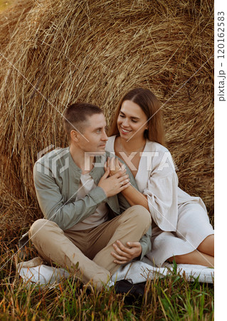 A couple is sitting on the grass next to a hay bale 120162583