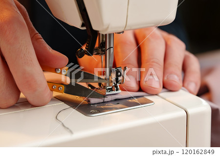 Hands sewing fabric using a sewing machine on a wooden table in a well-lit workspace Hands sewing fabric using a sewing machine on a wooden table in a well-lit workspace 120162849