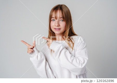 Young girl happily pointing against a neutral background while wearing a cozy white sweatshirt 120162896