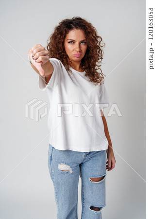 A woman with curly hair shows dislike with thumbs down against a neutral background A woman with curly hair shows dislike with thumbs down against a neutral background 120163058