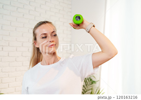 A woman exercises with green dumbbells at home in a bright room 120163216
