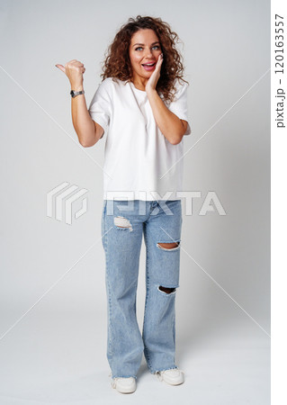 A young woman with curly hair playfully pointing finger while standing against a gray background 120163557