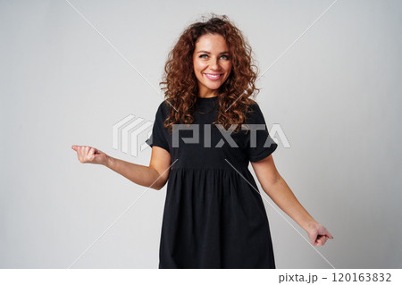 A young woman stands confidently in a black dress, showcasing a playful pose against a plain background 120163832