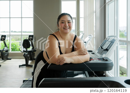 A plus-size woman stand on a gym treadmill, resting or preparing to exercise. 120164119