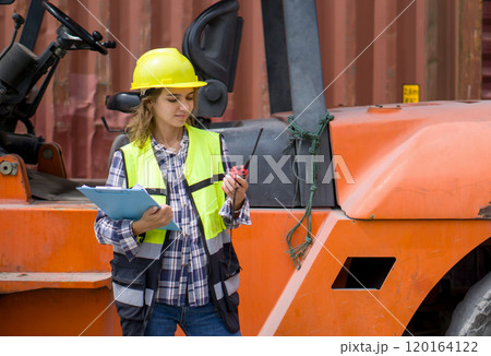 Young caucasian woman with yellow hardhat and safety vest stand in front of forklift while holding walkie talkie and clipboard. There are container in the work area. 120164122