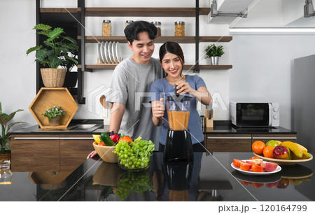 Young asian woman using blending machine mixing fruit and vegetable in to smoothie. Her boyfriend stand smiling behind. The kitchen counter full of various kinds of fruit and vegetable. Young asian woman using blending machine mixing fruit and vegetable in to smoothie. Her boyfriend stand smiling behind. The kitchen counter full of various kinds of fruit and vegetable. 120164799
