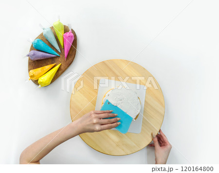 Closeup hand holding plastic cake cream spatula, spread the top of the cake with white cream, creating a decorative pattern on the top of a round cake on a wooden turntable or cake stand. Top view 120164800