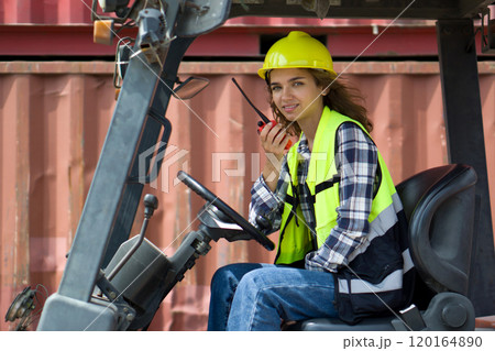 Young caucasian woman dressed in plaid pattern shirt, safety vest and yellow hardhat holding walkie talkie while sitting in forklift. There are container in the work area. Young caucasian woman dressed in plaid pattern shirt, safety vest and yellow hardhat holding walkie talkie while sitting in forklift. There are container in the work area. 120164890