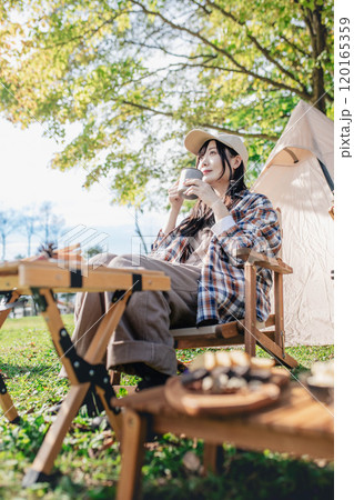 キャンプ場で椅子に座り飲み物を飲みながら休憩する女性 キャンプ場で椅子に座り飲み物を飲みながら休憩する女性 120165359