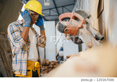 Construction worker in a yellow helmet using a chop saw to cut wood, with a vacuum cleaner collecting wood chips. woodworking, precision cutting, and construction sites, National Carpenters Day 120167086