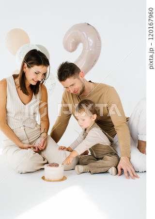 Young family with a child is sitting on the floor in front of a birthday cake in honor of the second birthday with balloons and the number two 120167960