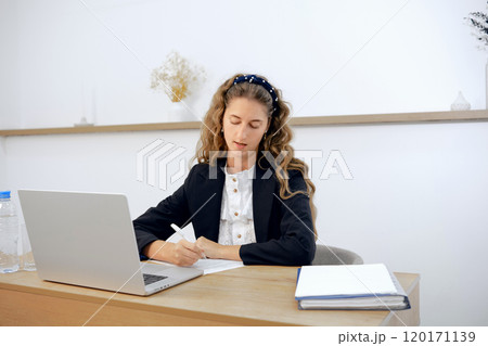 Focused young woman working on a project at a modern office desk during the daytime Focused young woman working on a project at a modern office desk during the daytime 120171139