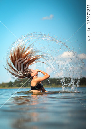 A young woman swimming in the clear blue ocean, her hair flowing in the water as she splashes around. She appears to be enjoying a refreshing summer day, with waves gently splashing around her 120171258