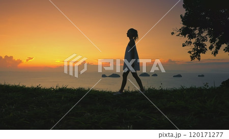 Woman gracefully walks grass hill, sunset sky in background. Female enjoy aerial view on tropical island. Girl silhouette against colorful orange sky. Outdoor lifestyle travel, summer holiday vacation Woman gracefully walks grass hill, sunset sky in background. Female enjoy aerial view on tropical island. Girl silhouette against colorful orange sky. Outdoor lifestyle travel, summer holiday vacation 120171277