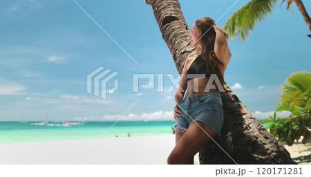 Young woman is enjoying her tropical vacation, relaxing on a white sand beach, leaning against a palm tree, with turquoise water and blue sky in the background 120171281
