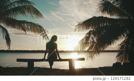 Silhouette of a young woman relaxing on a bench between two palm trees on a tropical island, enjoying a beautiful sunset over the water Silhouette of a young woman relaxing on a bench between two palm trees on a tropical island, enjoying a beautiful sunset over the water 120171282