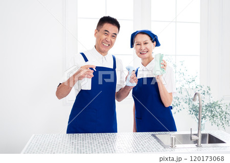青色のエプロンを付けキッチンに並んで立つ、白いポロシャツのシニアのハウスキーパー（男女、夫婦） 120173068