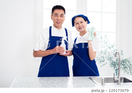 青色のエプロンを付けキッチンに並んで立つ、白いポロシャツのシニアのハウスキーパー(男女、夫婦) 青色のエプロンを付けキッチンに並んで立つ、白いポロシャツのシニアのハウスキーパー(男女、夫婦) 120173079