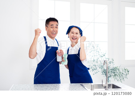青色のエプロンを付けキッチンに並んで立つ、白いポロシャツのシニアのハウスキーパー（男女、夫婦） 120173080