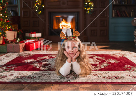 Merry Christmas. Little girl smiling near Christmas tree in classical dark interior. Young happy kid wearing deer horns in living room with fireplace Christmas tree gift boxes. Christmas eve at home 120174307