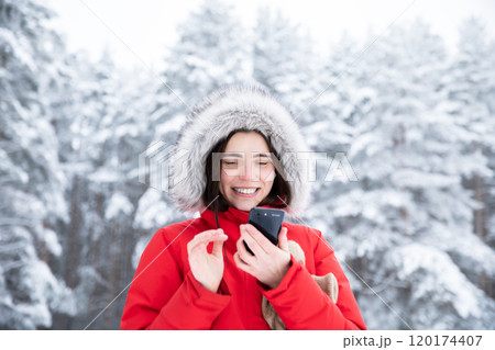 Beautiful woman in red hooded jacket, with phone in hands standing during day in winter snowy forest 120174407