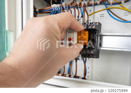 Close up of a hand inserting an electrical relay into a control panel with visible wiring and components. Close up of a hand inserting an electrical relay into a control panel with visible wiring and components. 120175894