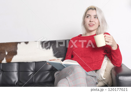 Woman in red sweater reading and holding a mug on a couch. 120177021