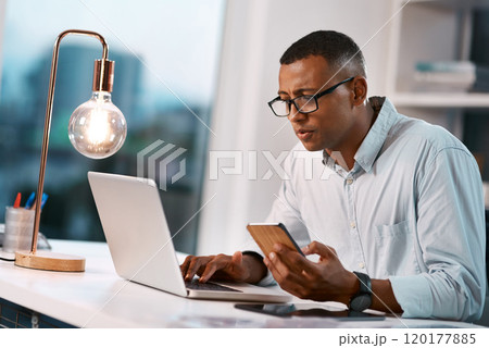 Hes a well connected man. Shot of a handsome young businessman using his laptop and cellphone while working late in his office. 120177885
