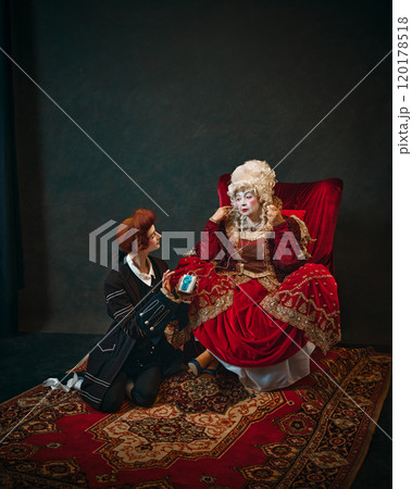 Humorous scene with elderly woman and young man. Medieval empress sitting in chair with young male servant offering her a music player. 120178518