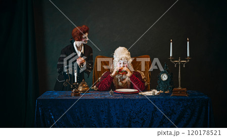 Young man, royal servant in wig, standing next to elderly woman, empress eating hamburger with passion, against dark vintage background. 120178521