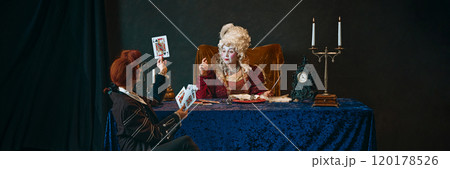 Elderly woman with whimsical makeup, medieval empress playing cards with young man, court favorite, set against vintage dark background. Elderly woman with whimsical makeup, medieval empress playing cards with young man, court favorite, set against vintage dark background. 120178526