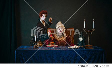 Young man, court servant serving hamburger with ketchup to serious elderly royal woman, empress sitting at table. 120178576