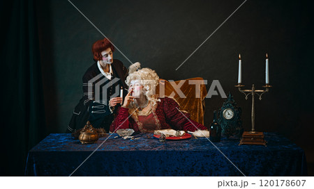 Royal empress, adorned in baroque gown, holding cigarette as her loyal favorite standing beside with candle. Royal empress, adorned in baroque gown, holding cigarette as her loyal favorite standing beside with candle. 120178607