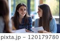 Two young women conversing and exchanging papers during a discussion 120179780