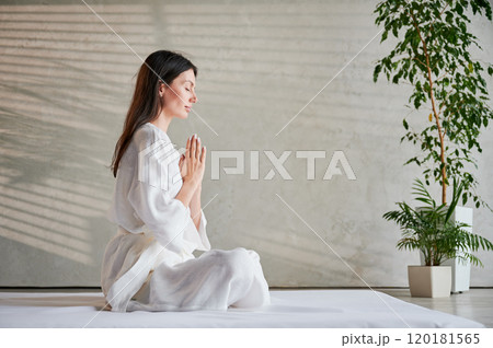 Beautiful woman in white dress sitting on mat in lotus position before Thai massage, practicing yoga, showing namaste sign. 120181565