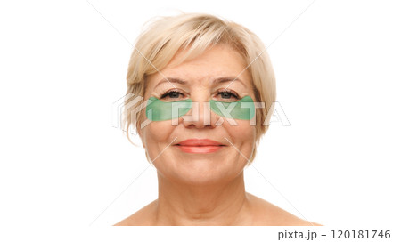 Close-up shot of senior blonde woman with green under-eye patches, looking directly at camera with a gentle smile against white studio background. 120181746