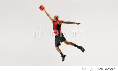 Young man in athletic clothing, wearing tank top and shorts, jumps with basketball toward hoop against grey studio background. Young man in athletic clothing, wearing tank top and shorts, jumps with basketball toward hoop against grey studio background. 120182597