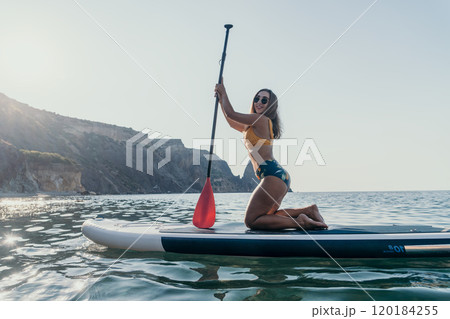 Woman Paddleboarding Ocean Scenic - Woman kneeling on a paddleboard in the ocean, holding the paddle. The scenery behind her is mountainous. 120184255
