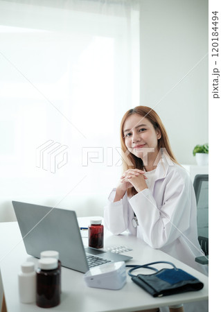 A woman in a white lab coat sits at a desk with a laptop and a bottle of pills 120184494