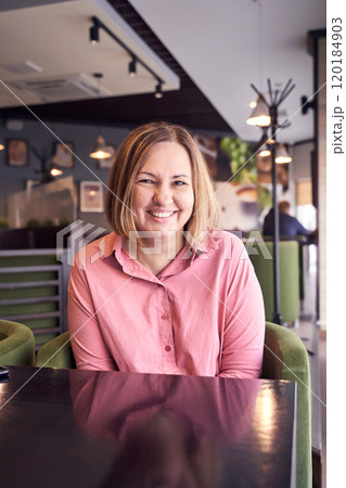 Smiling woman in pink shirt relaxing at modern cafe with cozy interior and green 120184903