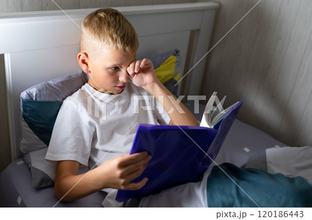 fair-haired teenage boy reads a book before bed and rubs his eyes, wants to sleep fair-haired teenage boy reads a book before bed and rubs his eyes, wants to sleep 120186443