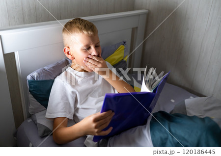 fair-haired teenage boy reads a book before bed and yawns 120186445