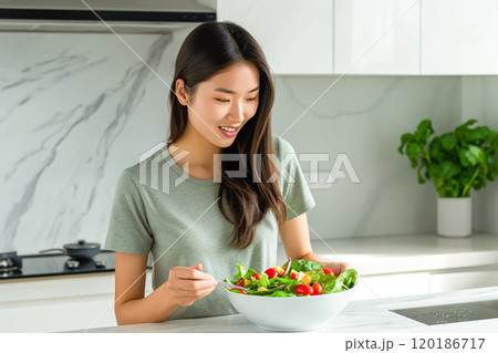 Asian woman eating salad in a light kitchen, healthy eating concept 120186717