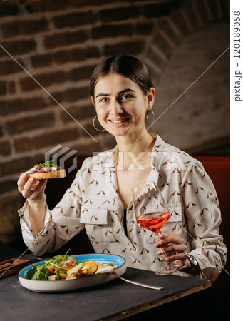 Smiling woman enjoying a gourmet brunch with toast and a glass of rose wine in a cozy restaurant Smiling woman enjoying a gourmet brunch with toast and a glass of rose wine in a cozy restaurant 120189058