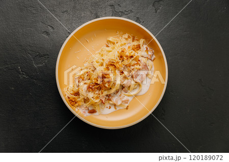 Creamy tagliatelle pasta with crispy bacon and fried onions in a beige bowl on a dark background 120189072