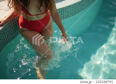 A woman in a vibrant red bikini is playfully splashing water while sitting on the edge of a pool. The scene captures a moment of relaxation and joy, highlighting the shimmering blue water and the 120189745