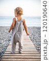 Beautiful toddler in summer grey jumpsuit stands alone on path leading to the seashore on warm day. 120190640