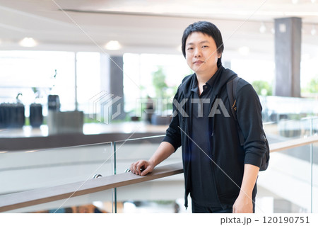 Standing next to the terrace barricade in a modern building, an Asian man exudes confidence in his stylish black attire. Standing next to the terrace barricade in a modern building, an Asian man exudes confidence in his stylish black attire. 120190751